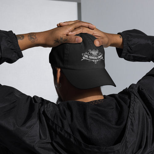 Person wearing a black cap with The Crystal Cowboy emblem embroidered on the back, hands clasped behind their head against a neutral background.